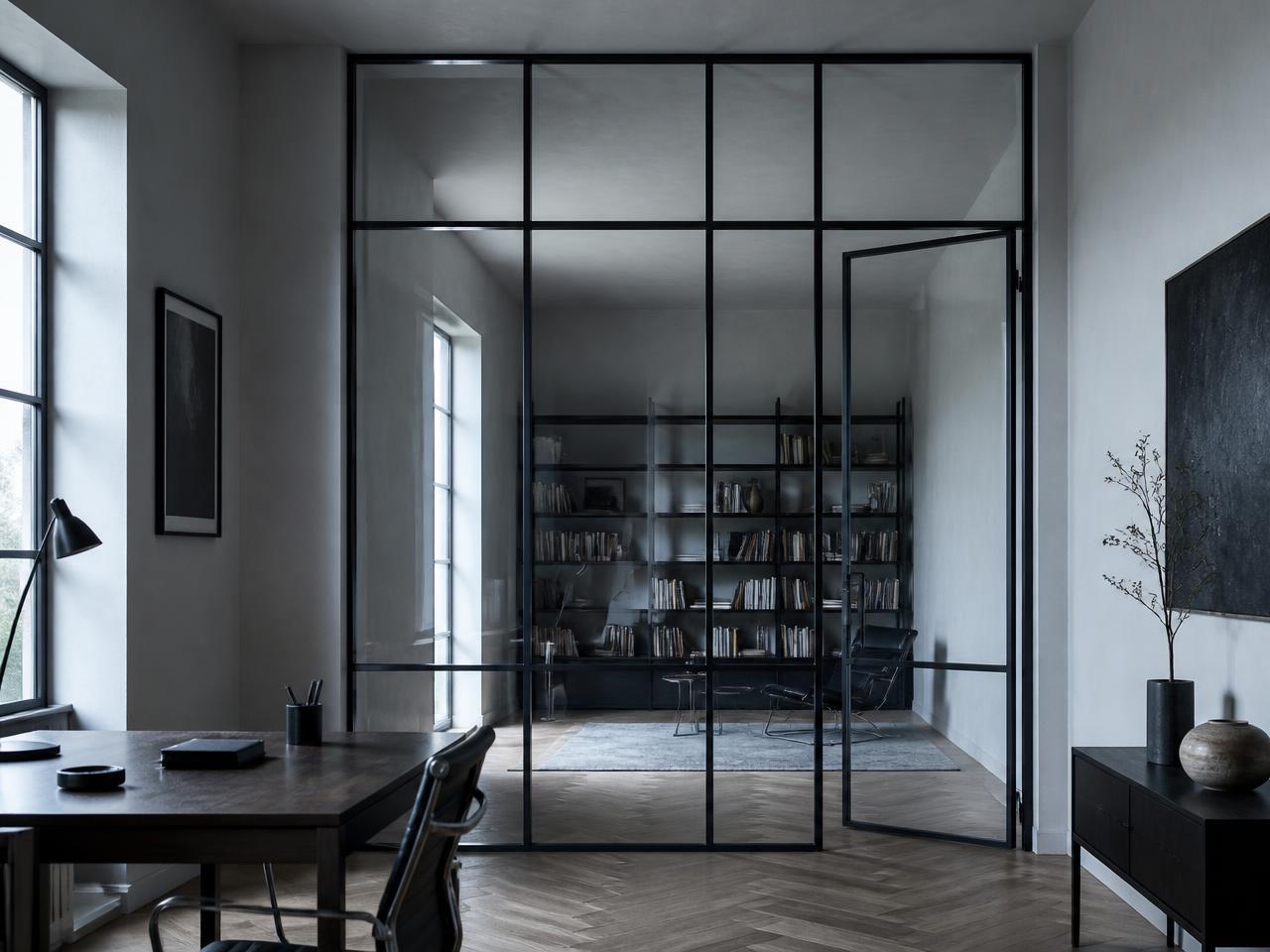 Slim black steel and glass interior partition dividing a minimal study from a library beyond