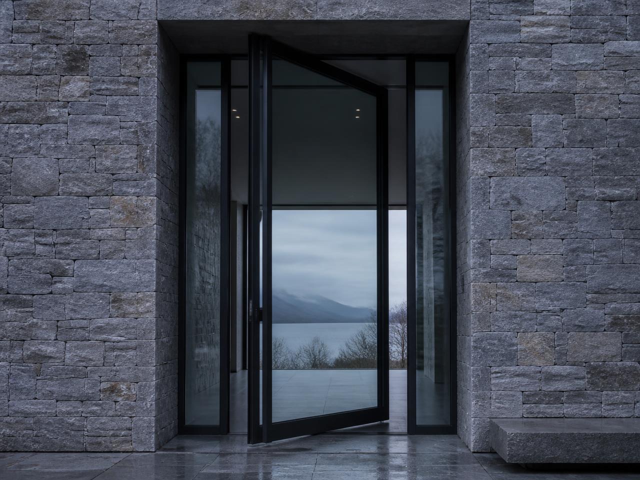 Tall black steel pivot entry door with narrow sightlines set into a stone facade